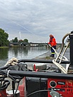 Im Vordergrund eine Motorpumpe, im Hintergrund ein THW-Helfer der mit einem Strahlrohr ins Wasser spritzt.